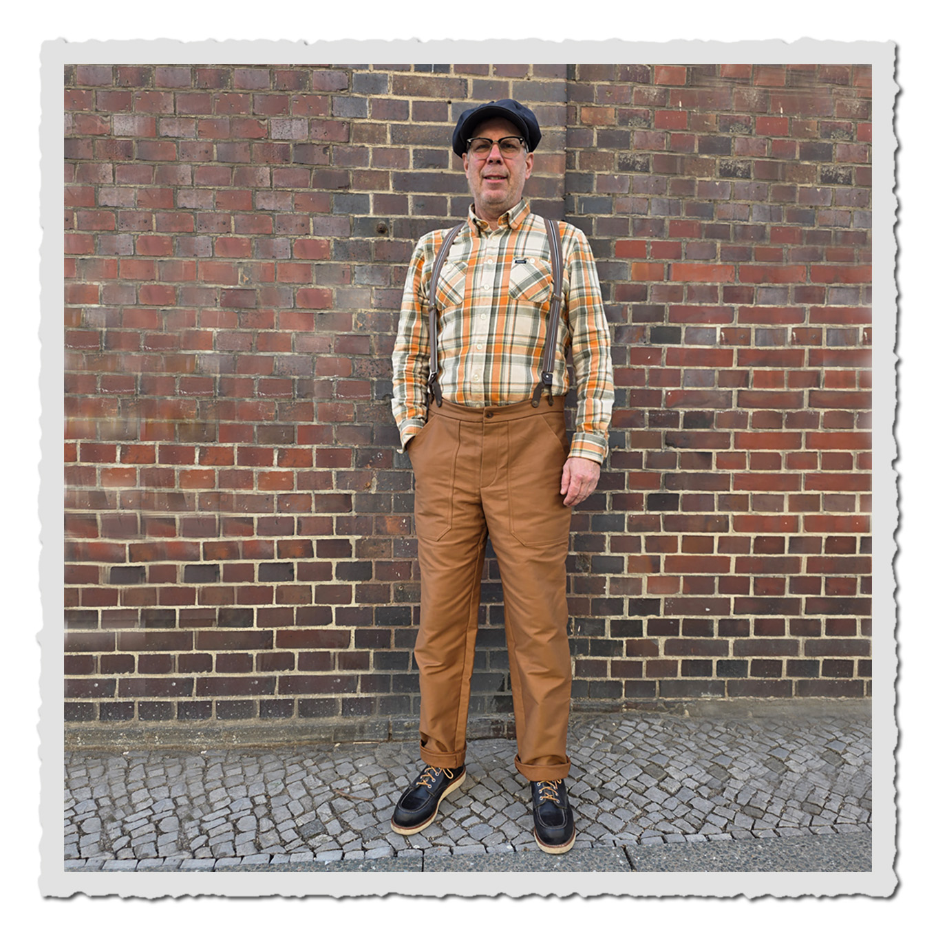 Ganzkörperfoto: Mann mit Hosenträgern und brauner Hose, vor roter Ziegelwand im Freien.